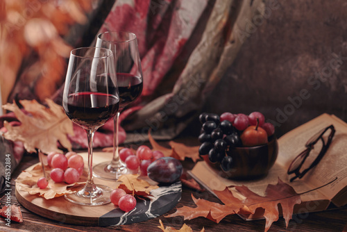 Autumn still life with two glasses of red wine, grapes, book and dry leaves in rustic style on dark wooden background. Romantic sweater weather, wine tasting concept with copy space