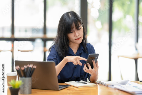 Smiling Asian businesswoman talking on the phone while working with laptop in office