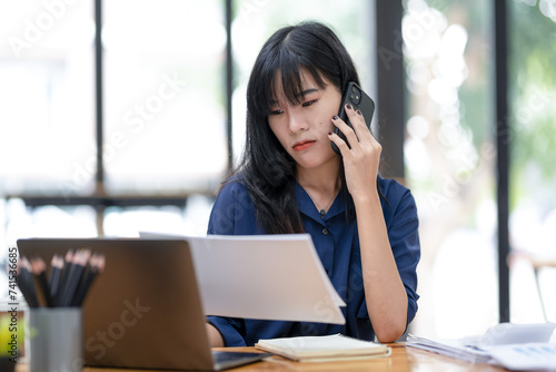 Smiling Asian businesswoman talking on the phone while working with laptop in office