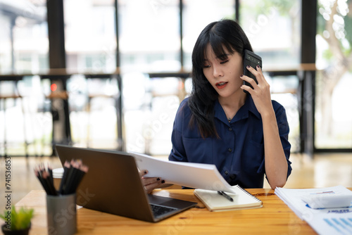 Smiling Asian businesswoman talking on the phone while working with laptop in office