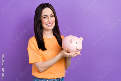 Photo of young smiling woman employee holding piggy bank finally collected money for dream journey isolated over violet color background