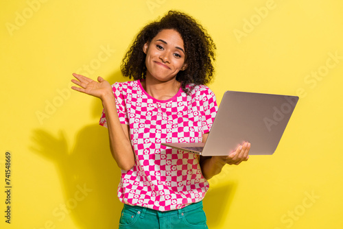 Portrait of attractive clueless person hold wireless netbook raise hand shrug shoulder isolated on yellow color background photo