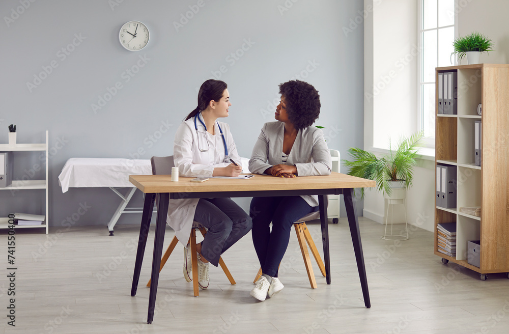 Female doctor and patient communicating in medical room at clinic. Young physician sitting at desk together with African American woman, writing out prescription and explaining how to take medicine Female doctor and patient communicating in medical room at clinic. Young physician sitting at desk together with African American woman, writing out prescription and explaining how to take medicine