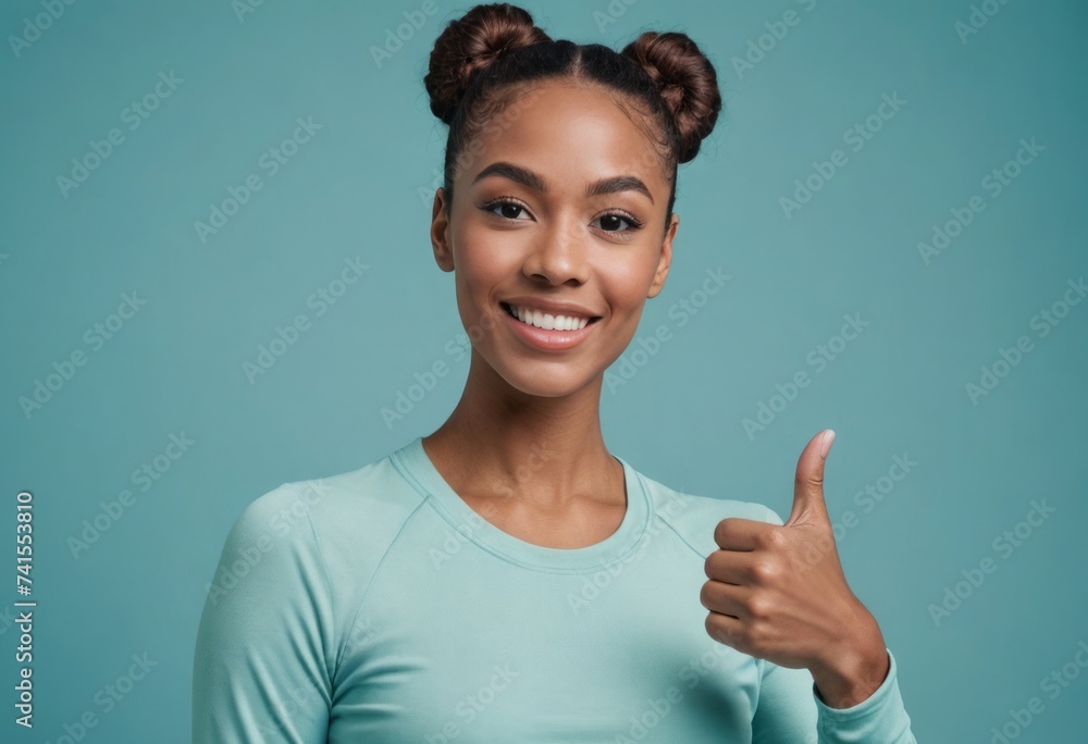 A woman with hair in buns, making a friendly thumbs up gesture in active wear. Studio shot with a blue background. - obrazy, fototapety, plakaty 