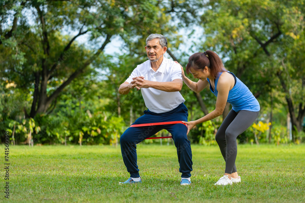 Senior asian man is using rubber band to build up his leg muscle strength while his daughter is cheering up in the public park for elder longevity exercise and outdoor workout Senior asian man is using rubber band to build up his leg muscle strength while his daughter is cheering up in the public park for elder longevity exercise and outdoor workout