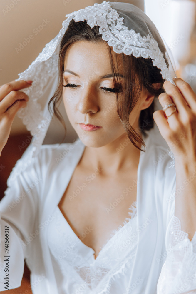Fototapeta premium portrait of an incredibly beautiful girl bride in a white robe in the bedroom, the bride poses holding her veil in her hands and covers herself with it.