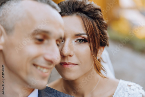 Portrait of a smiling bride with a bouquet of flowers hugging her husband while looking at the camera. Beautiful wedding celebration. Wedding hairstyle with a veil. Couple in love.