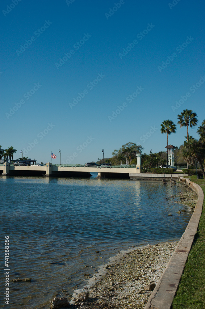 View north from Sunset Park in Saint Petersburg, FL with leading line of seawall with grass and palm trees on right. Bridge and other trees in middle and left. Late afternoon sun across bay water.