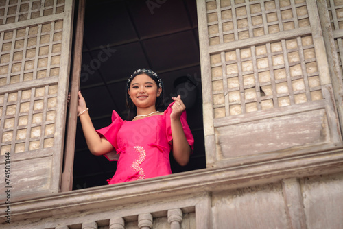 A poised young lady in a vibrant pink Filipiniana gown smiling by the window of an ancestral house, making a heart finger gesture. photo