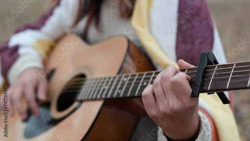 Close up of girl playing the guitar with capo outdoors on autumn nature. Girl covered in warm plaid play acoustic guitar outdoors