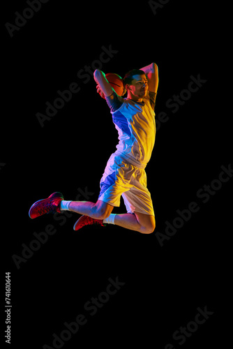 Athlete man, basketball player, in motion of powerful basketball shot against black studio background in mixed neon light. Concept of professional sport, energy, strength and power, match, tournament.