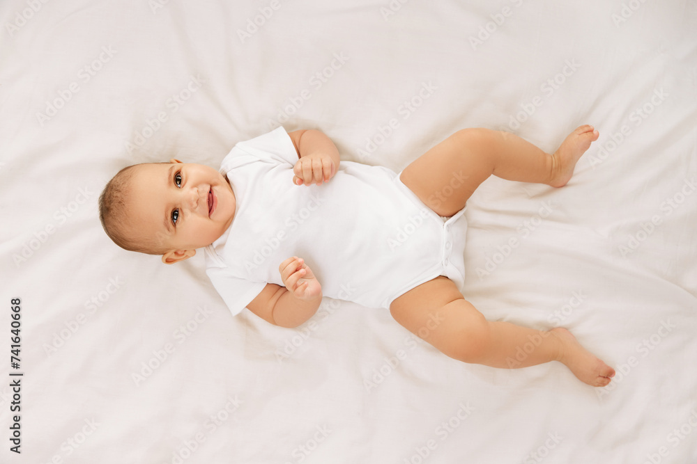 Happy, smiling child, baby, adorable girl in white onesie lying on bed and lookin upwards against white background. Concept of childhood, family, care, motherhood, infancy, heath Happy, smiling child, baby, adorable girl in white onesie lying on bed and lookin upwards against white background. Concept of childhood, family, care, motherhood, infancy, heath