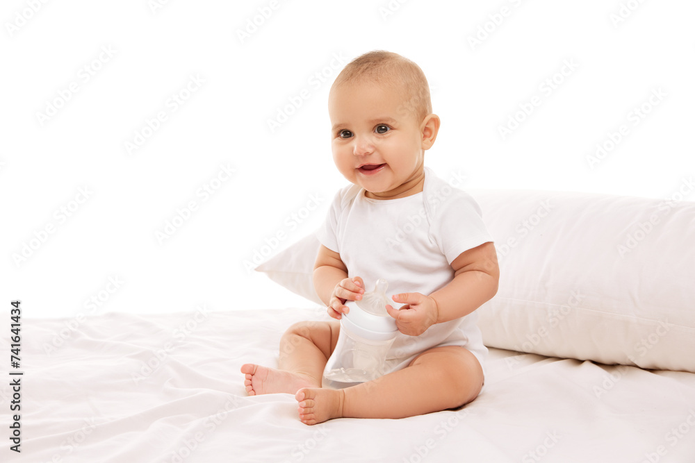 Little baby child, girl, adorable infant in white onesie sitting on bed with pillow and smiling against white background. Concept of childhood, family, care, motherhood, infancy, heath Little baby child, girl, adorable infant in white onesie sitting on bed with pillow and smiling against white background. Concept of childhood, family, care, motherhood, infancy, heath