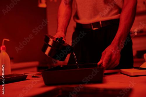 cropped photographer pours developer into a tray for film processing in darkroom with red light photo
