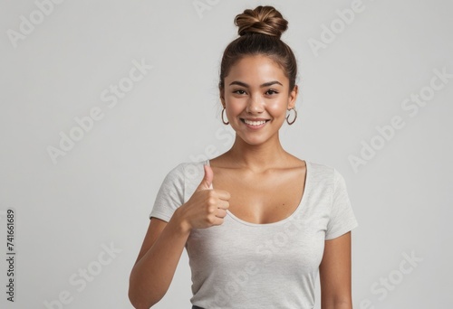 A young woman with a top knot hairstyle smiles brightly, giving a thumbs up in a casual white top. © kotlyarn