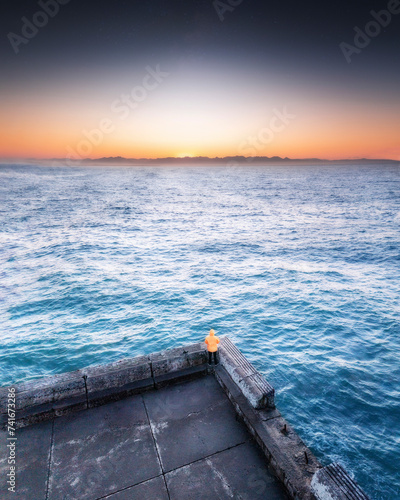 Aerial view of a man in an orange jacket, standing on the edge of a jetty looking out towards the sunset with mountains on the horizon, Kalk Bay, Western Cape, South Africa. photo