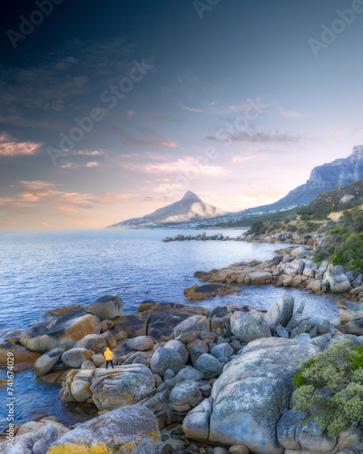 Aerial view of a man in an orange jacket standing on a boulder overlooking a calm ocean and lions head mountain in the background with whispy clouds at sunrise, Cape Town, Western Cape, South Africa. photo