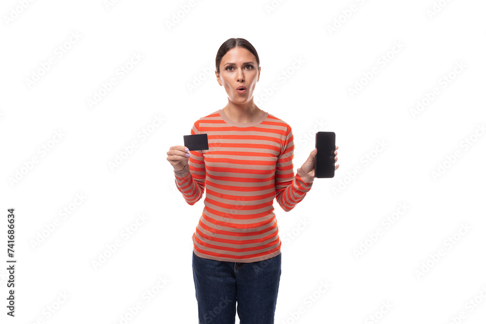 A young pretty brunette woman dressed in a striped orange blouse with surprise shows a plastic card and a smartphone A young pretty brunette woman dressed in a striped orange blouse with surprise shows a plastic card and a smartphone