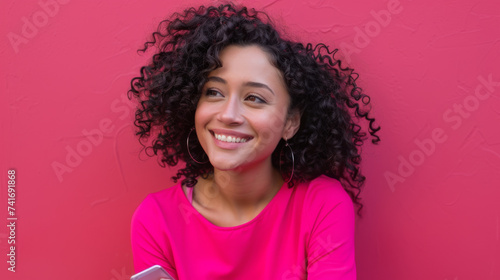 cheerful young woman with curly hair is holding a smartphone and looking away with a smile against a vibrant pink background. © MP Studio