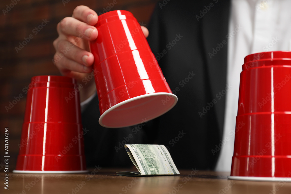 Shell game. Man showing dollar banknote under cup at wooden table, closeup - obrazy, fototapety, plakaty 