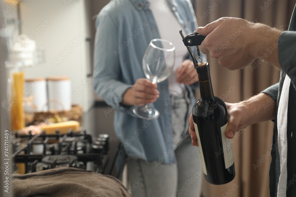Man opening wine bottle with corkscrew while woman holding wineglass indoors, closeup