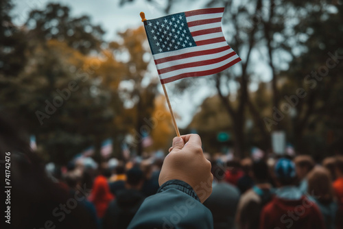 Close-up of a hand holding an American flag, with a crowd of people in the background, symbolizing individual and collective pride on Flag Day