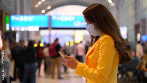 Woman in airport terminal, adorned in vibrant yellow jacket and medical mask, using smartphone. She checks updates on her travel plans, possibly sending messages or notifications to loved ones. photo