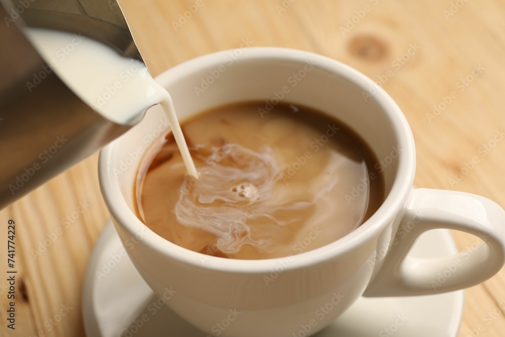 Pouring milk into cup with coffee at light wooden table, closeup Pouring milk into cup with coffee at light wooden table, closeup