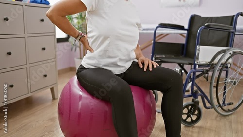 Mature woman exercises on fitness ball in rehab clinic with wheelchair in background photo