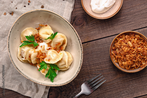 A plate of pelmeni, Russian meat dumplings, garnished with parsley and topped with sour cream and crispy onions, served with a fork on a wooden table.