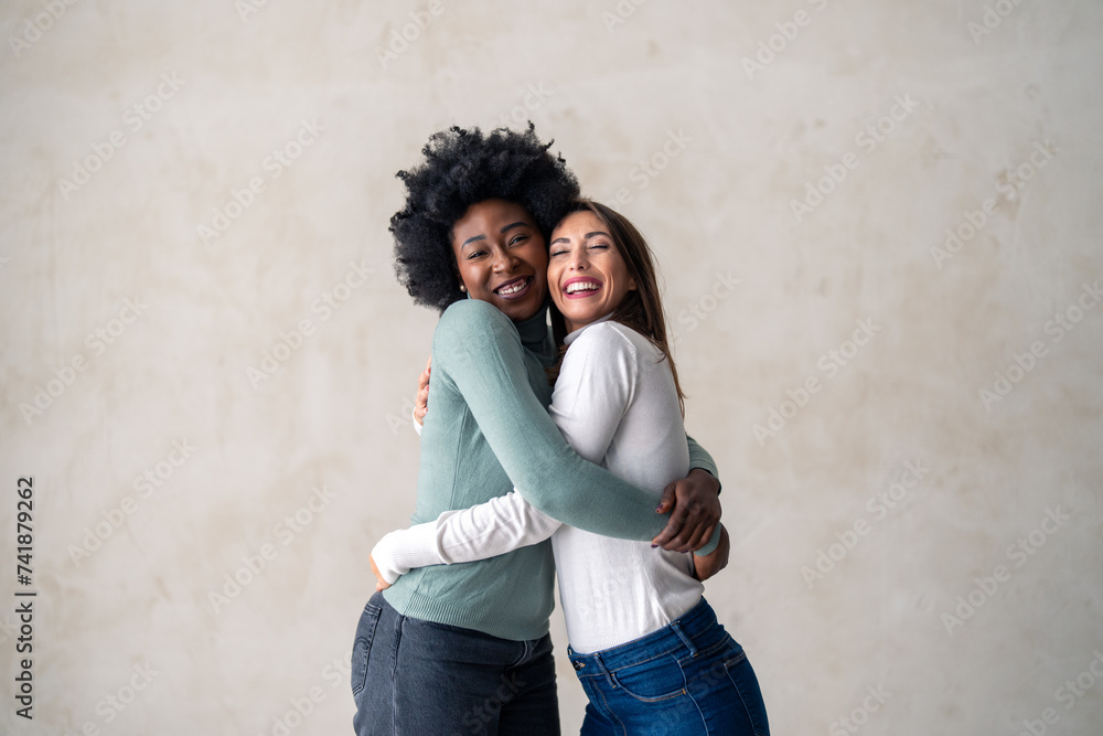 Two best friends smiling while embracing each other in a studio. Happy young women enjoying themselves while standing against a background in a studio.