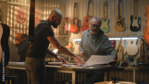 Two professional carpenters, craftsmen inspect wooden guitar body blank, discuss process of making musical instrument in modern workshop. Colleague works in background. Handmade and entrepreneurship.