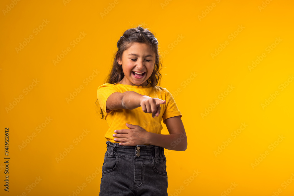 Kid girl mocking and teasing at someone showing finger at camera and holding stomach over yellow background. Bulling concept Kid girl mocking and teasing at someone showing finger at camera and holding stomach over yellow background. Bulling concept