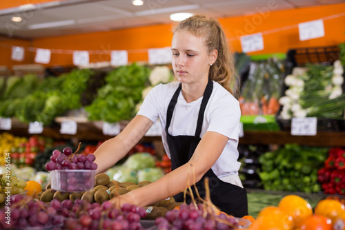 Fotografie Hardworking fifteen-year-old girl who works part-time in a store as a trainee saleswoman puts grapes on the counter, laid