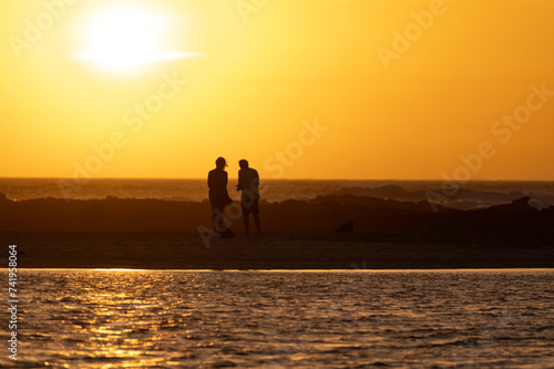 Man and woman staying and looking at sunset on ocean beach, orange sky, silhouettes of people on vacation © barmalini