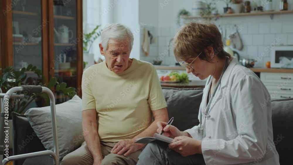 Female doctor in white coat sitting on couch, asking elderly man about his health and filling out medical record form during consultation at home Female doctor in white coat sitting on couch, asking elderly man about his health and filling out medical record form during consultation at home
