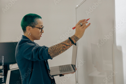 Focused tattooed business woman working on laptop and making notes on flipchart on office background