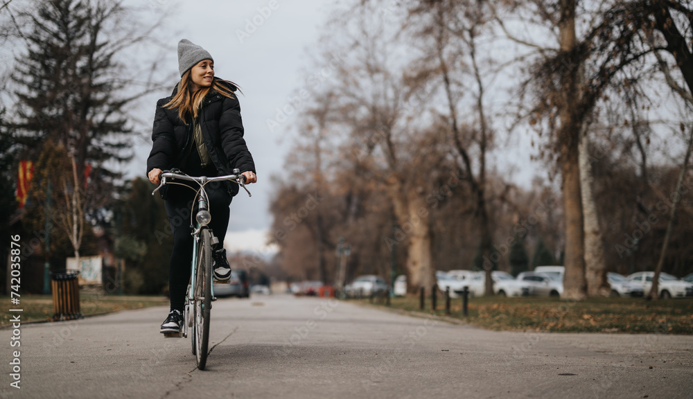 An active young woman wearing a beanie and jacket smiles while cycling down a tree-lined street on a cool fall day, embracing outdoor lifestyle.