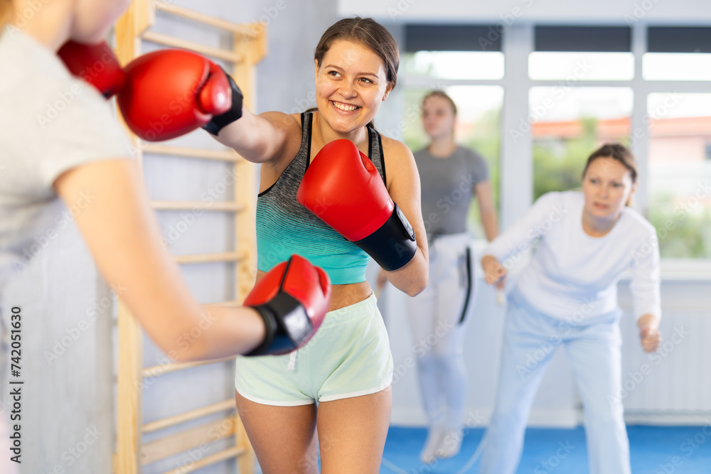Concentrated and cheerful adolescent girl in boxing gloves practicing punches in sparring during group self defence course supervised by female instructor in gym..