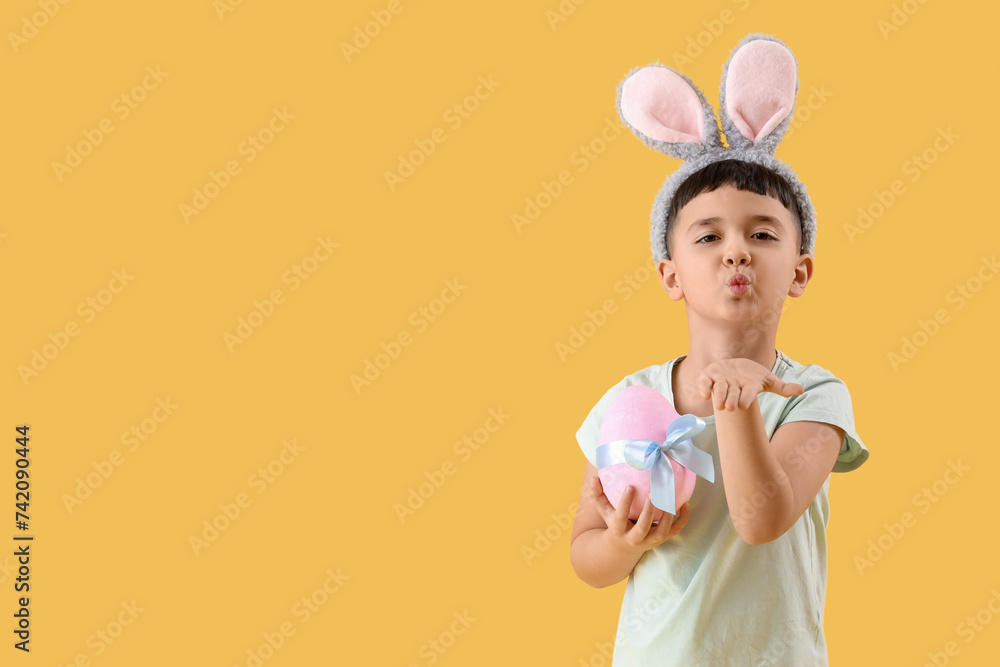 Cute little boy wearing bunny ears with Easter gift egg blowing kiss on yellow background Cute little boy wearing bunny ears with Easter gift egg blowing kiss on yellow background