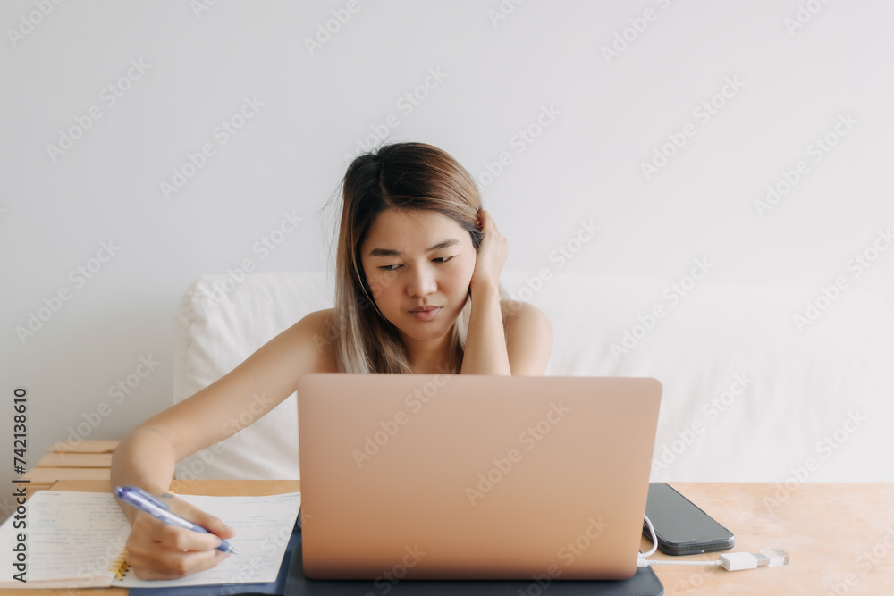 Serious freelance Asian woman is working online at home with computer laptop and taking notes in her apartment. Serious freelance Asian woman is working online at home with computer laptop and taking notes in her apartment.