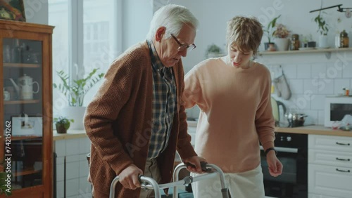 Young woman supporting elderly grandfather getting around the house with a walking frame, teaching him how to use mobility aid. Tilt-down shot