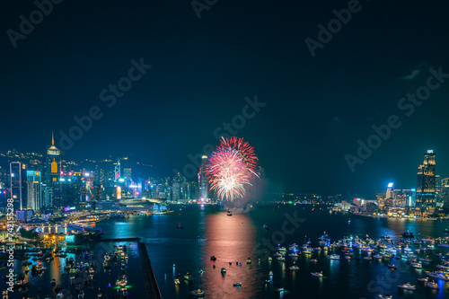 Fireworks burst brightly against the night sky above a city skyline, reflecting in the calm water below
