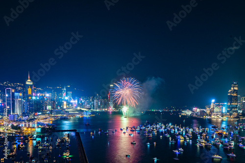 Fireworks burst brightly against the night sky above a city skyline, reflecting in the calm water below