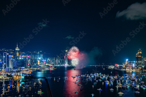 Fireworks burst brightly against the night sky above a city skyline, reflecting in the calm water below