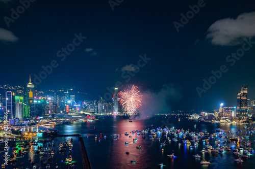 Fireworks burst brightly against the night sky above a city skyline, reflecting in the calm water below