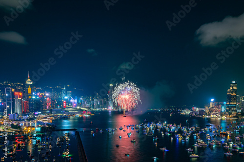 Fireworks burst brightly against the night sky above a city skyline, reflecting in the calm water below