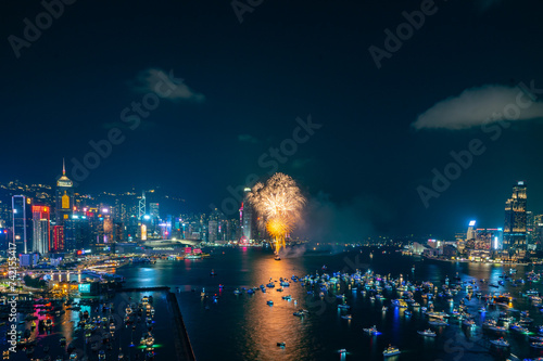 Fireworks burst brightly against the night sky above a city skyline, reflecting in the calm water below