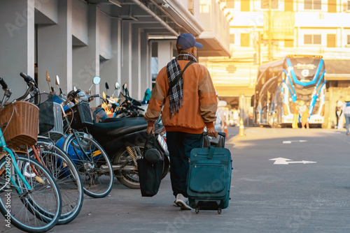 Back view of elderly traveler man in casual clothes dragging suitcase . Senior man walking with his luggage on street in morning. Male tourist with luggage for vacation. Vacation and travel concept.