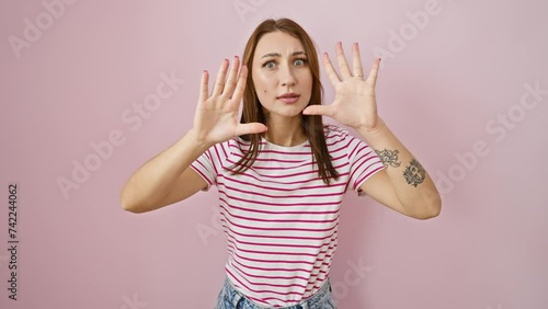 Angry, young brunette girl in striped tshirt expresses frustration with stop gesture, hands over pink isolated background